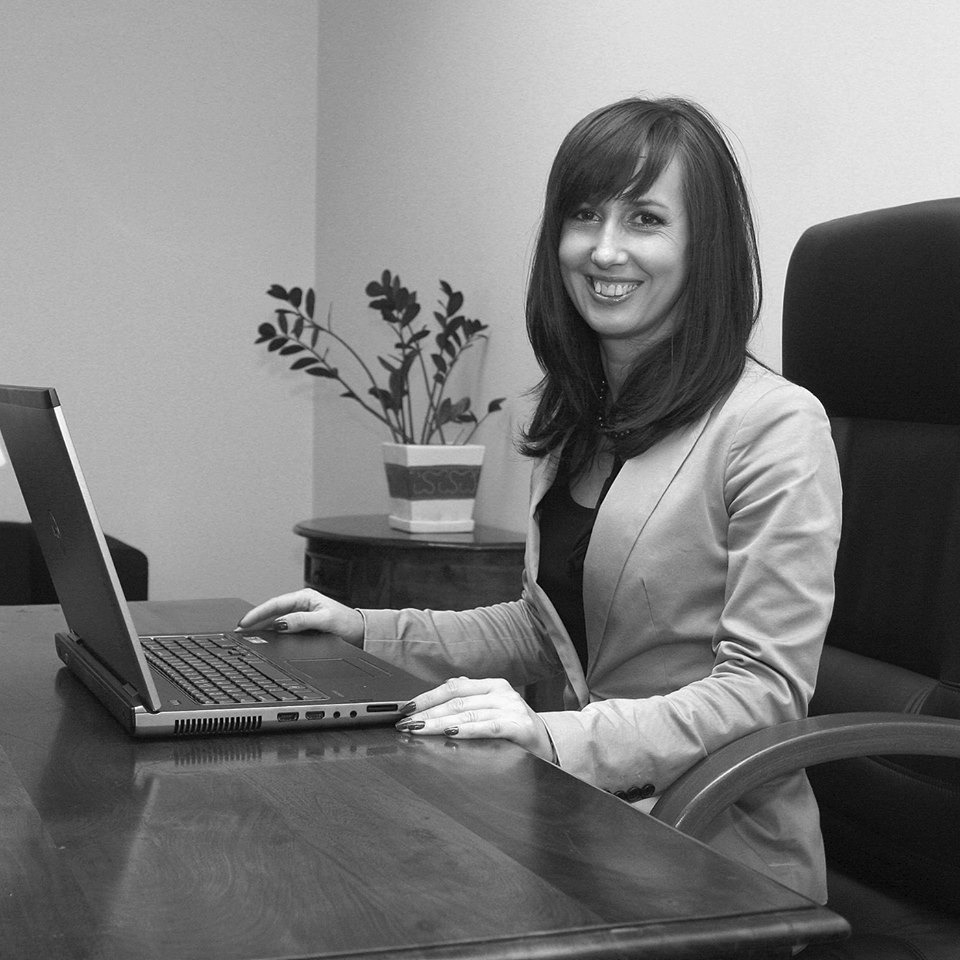 Portret uśmiechniętej kobiety w marynarce, pracującej na laptopie przy biurku w biurze, obok stoi roślina w ozdobnej doniczce.