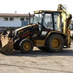 Żółta koparko-ładowarka Caterpillar 428C stoi na betonowym placu, z łyżką wypełnioną ziemią i widocznym budynkiem w tle.