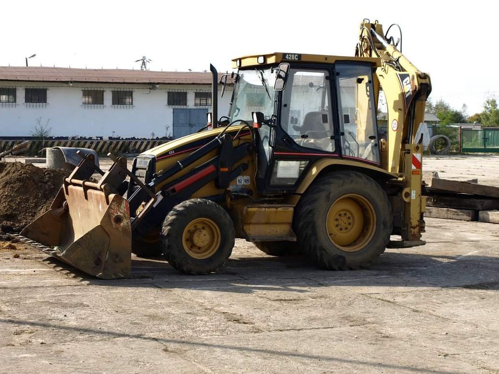Żółta koparko-ładowarka Caterpillar 428C stoi na betonowym placu, z łyżką wypełnioną ziemią i widocznym budynkiem w tle.