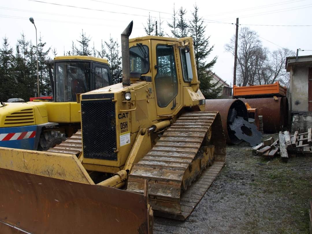 Żółty, używany spychacz CAT D5H na placu budowy z widocznymi śladami eksploatacji, obok walec drogowy i inne maszyny budowlane, w tle drzewa i budynek.
