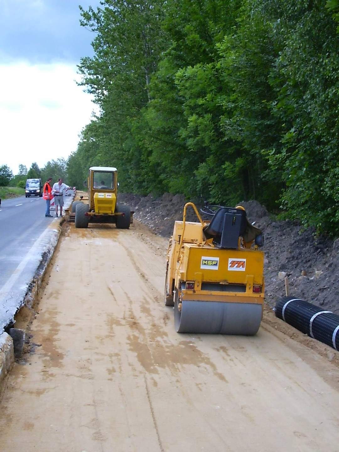Żółty walec drogowy ubija piasek na poboczu drogi, w tle widoczny drugi walec i dwóch pracowników w kamizelkach odblaskowych.
