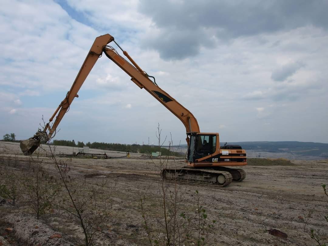 Pomarańczowa koparka CAT 320D z uniesionym ramieniem i łyżką na terenie budowy, widoczne rury i gruz, w tle pochmurne niebo.