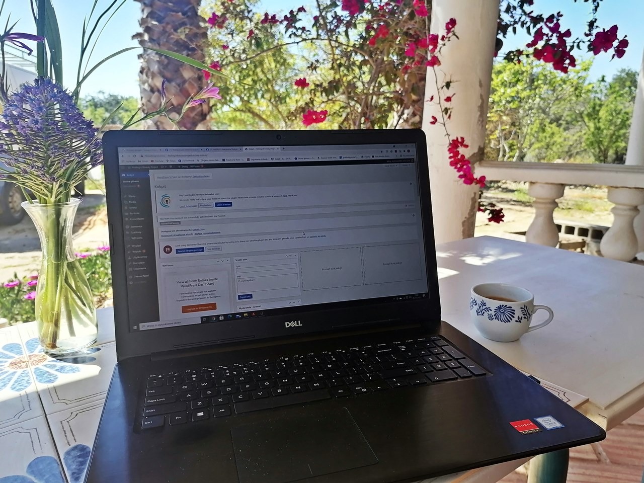 Laptop z otwartym panelem WordPress na stole na werandzie, obok filiżanka kawy i wazon z kwiatami, w tle bujna roślinność.