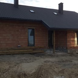 AB usługi budowlane - Surowy stan domu z czerwonej cegły z czarnym dachem i oknami. Widoczne deski i piasek przed budynkiem. Częściowo otwarte drzwi samochodu po lewej stronie.