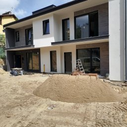 AB usługi budowlane - Nowoczesny, dwukondygnacyjny dom w trakcie wykończenia elewacji. Widoczny piasek, drabina i materiały budowlane. Część elewacji w kolorze białym, część w odcieniach szarości.