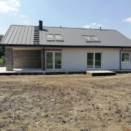 Nowoczesny dom jednorodzinny z szarą blachodachówką, trzema oknami dachowymi i drabinką kominiarską, fragment elewacji wykończony cegłą klinkierową, teren wokół domu jeszcze niezagospodarowany.