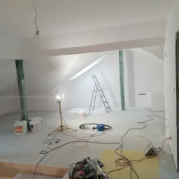 Poddasze po malowaniu na biało, widoczny sprzęt malarski, szlifierka do gładzi, drabina aluminiowa, wiadro z farbą, lampa halogenowa na trójnogu, zabezpieczone filarem drewniane, gniazdka elektryczne.