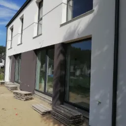 Dwukondygnacyjny dom z elewacją w kolorze białym i brązowym, z dużymi oknami i stertami drewnianych podestów przed nimi, widoczne przewody elektryczne wystające ze ściany.