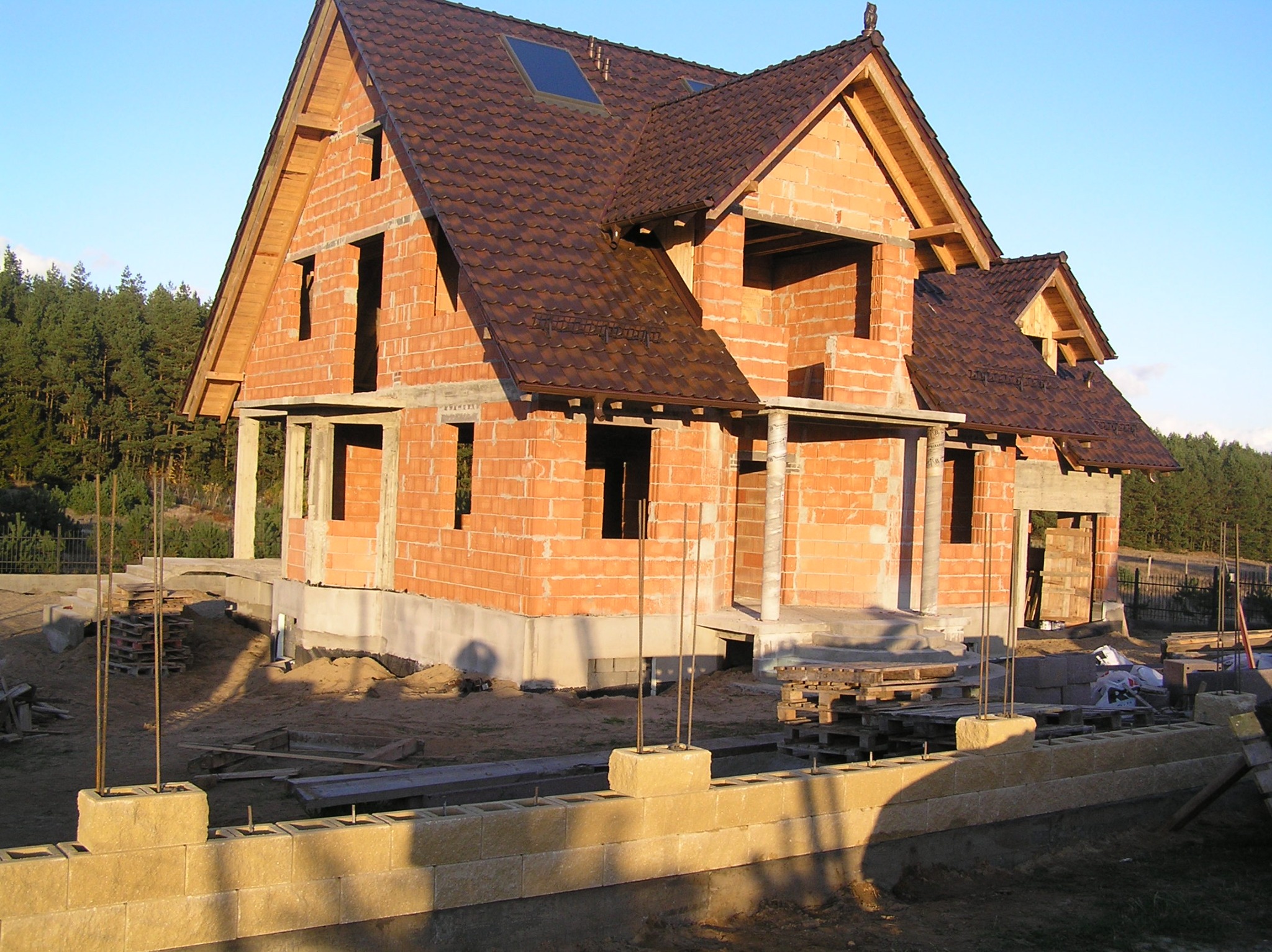 Surowy stan domu z czerwonej cegły z brązowym dachem, widoczne otwory okienne i drzwiowe, fundament z bloczków, teren budowy z paletami i zbrojeniami.