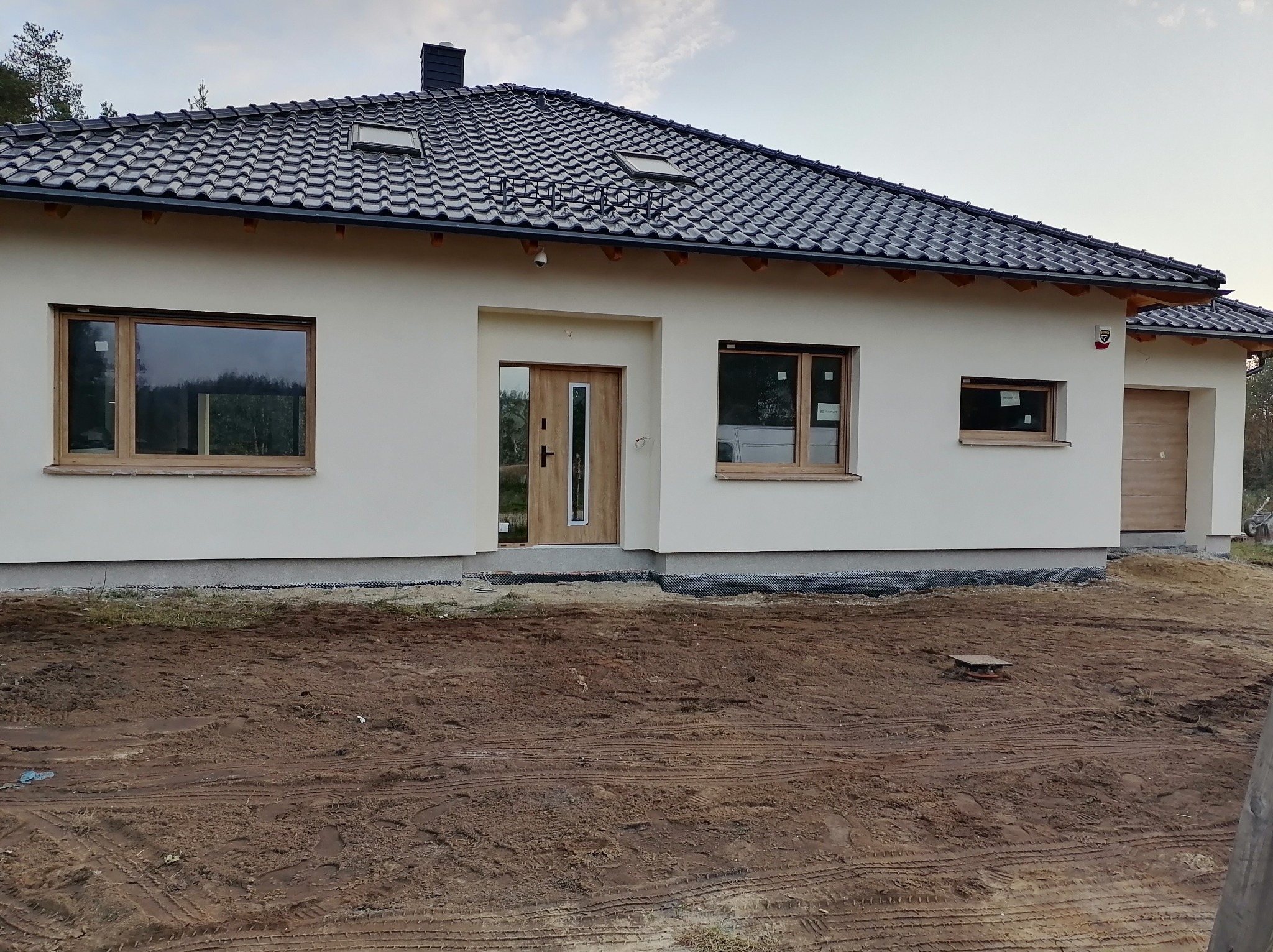 Nowoczesny dom jednorodzinny z jasną elewacją, drewnianymi oknami i ciemną dachówką. Widoczne detale wykończenia i przygotowany teren wokół budynku.