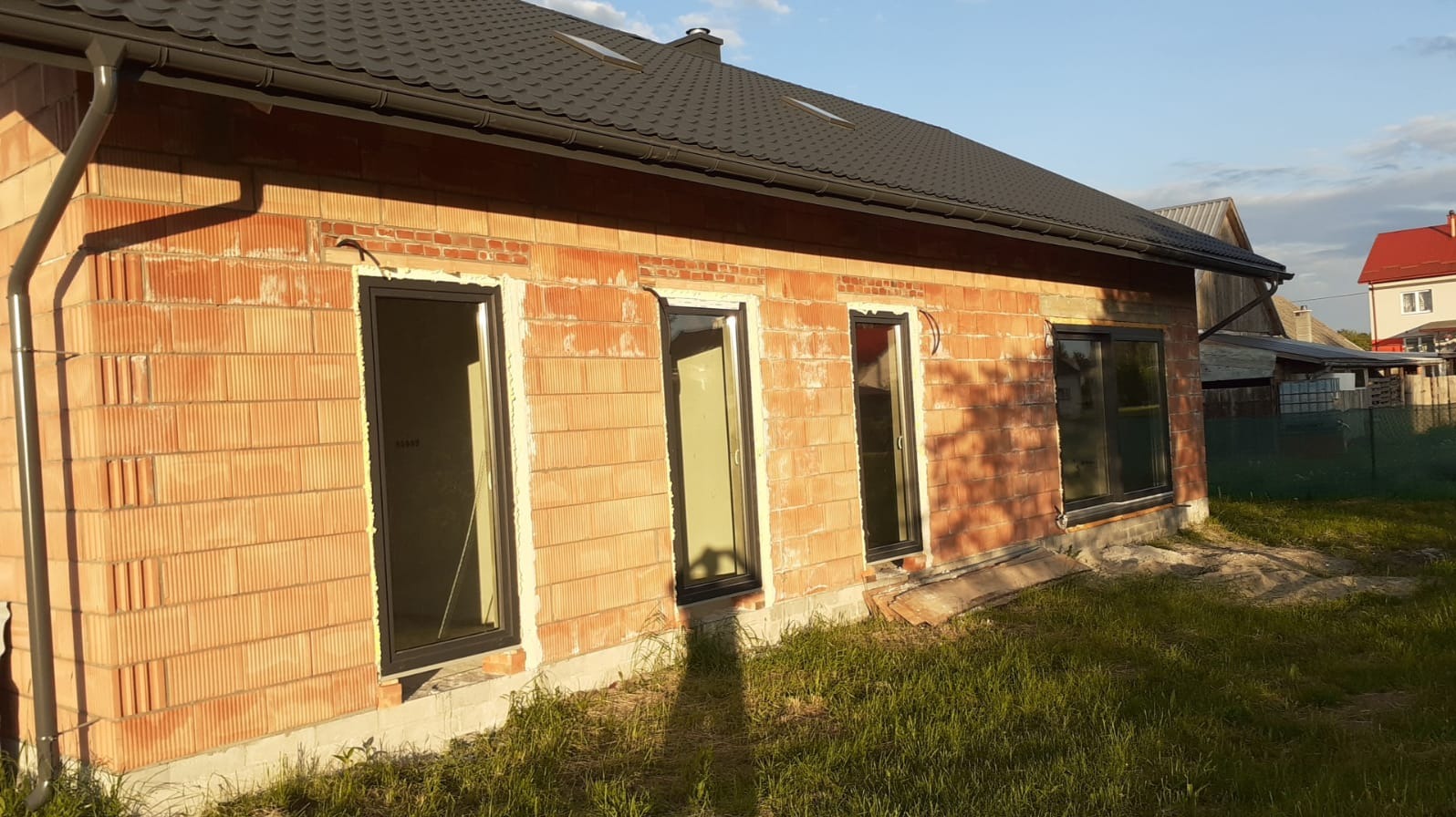 Surowy mur z pustaków ceramicznych z zamontowanymi oknami w czarnych ramach, widok z zewnątrz, w tle fragment dachu z rynnami i zieleń trawy.