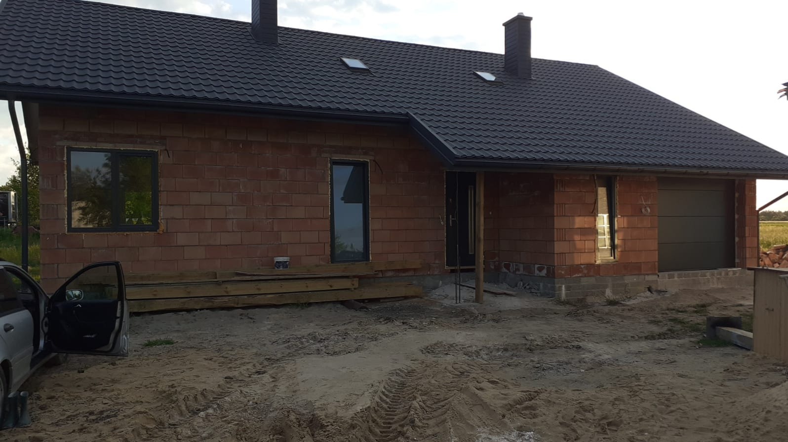 Surowy stan domu z czerwonej cegły z czarnym dachem i oknami. Widoczne deski i piasek przed budynkiem. Częściowo otwarte drzwi samochodu po lewej stronie.