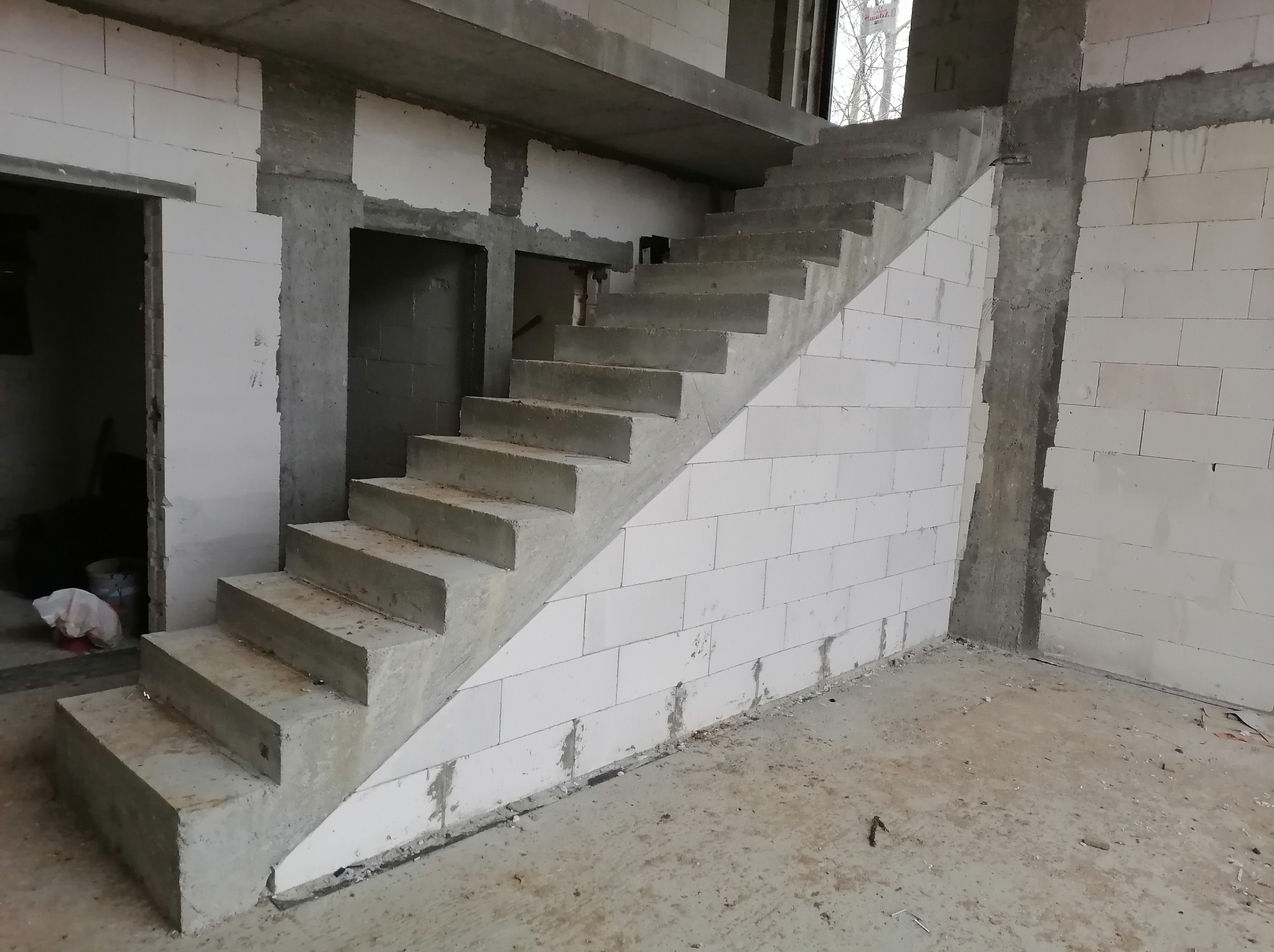 Surowe betonowe schody wewnątrz budynku w trakcie budowy, ściany z białych bloczków, widoczne otwory drzwiowe i okno. Stan surowy zamknięty.