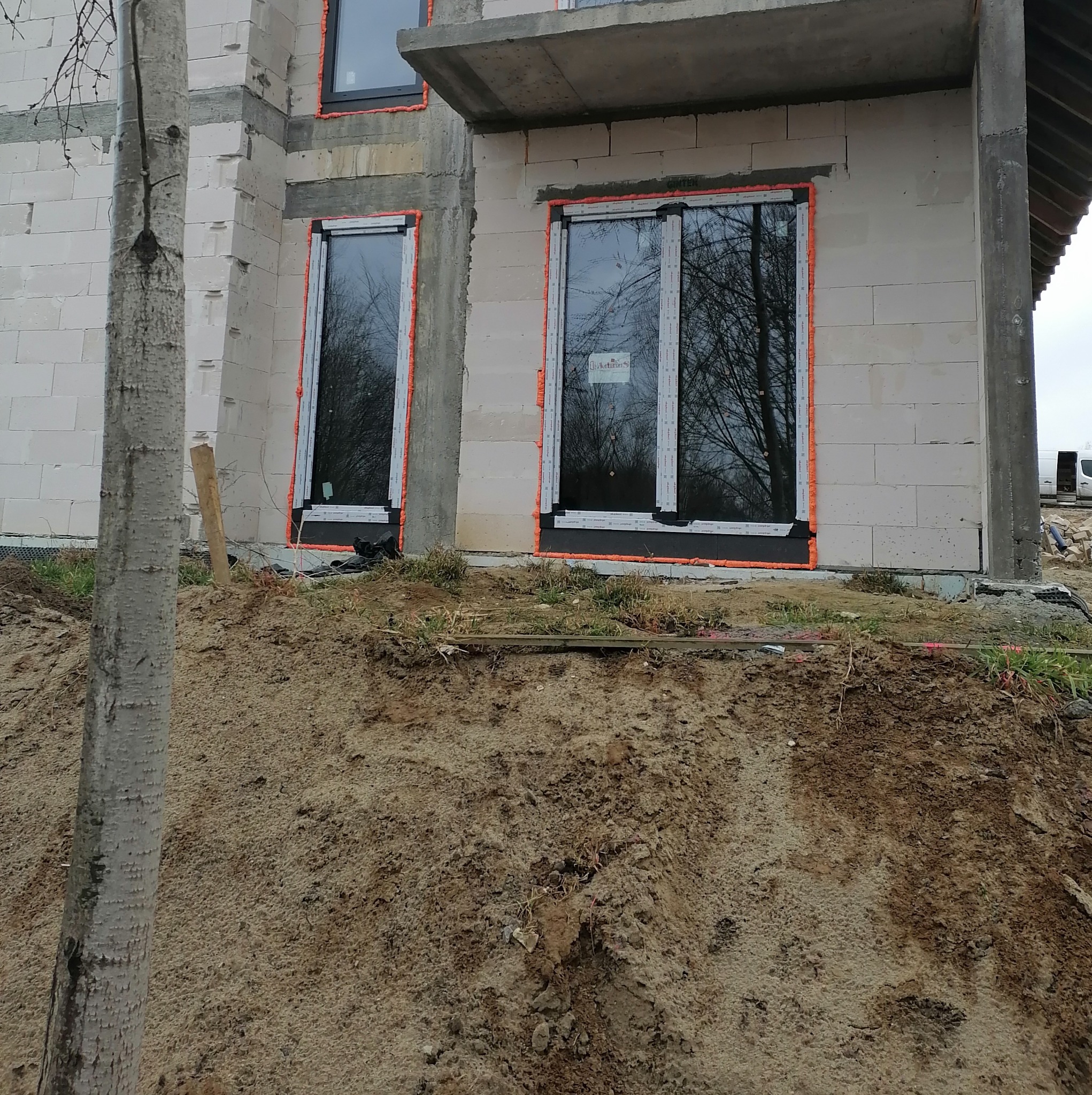 Budowa domu z betonu komórkowego: okna z pomarańczową pianką montażową, widoczny fragment elewacji i ziemia. Surowy stan, etap prac.