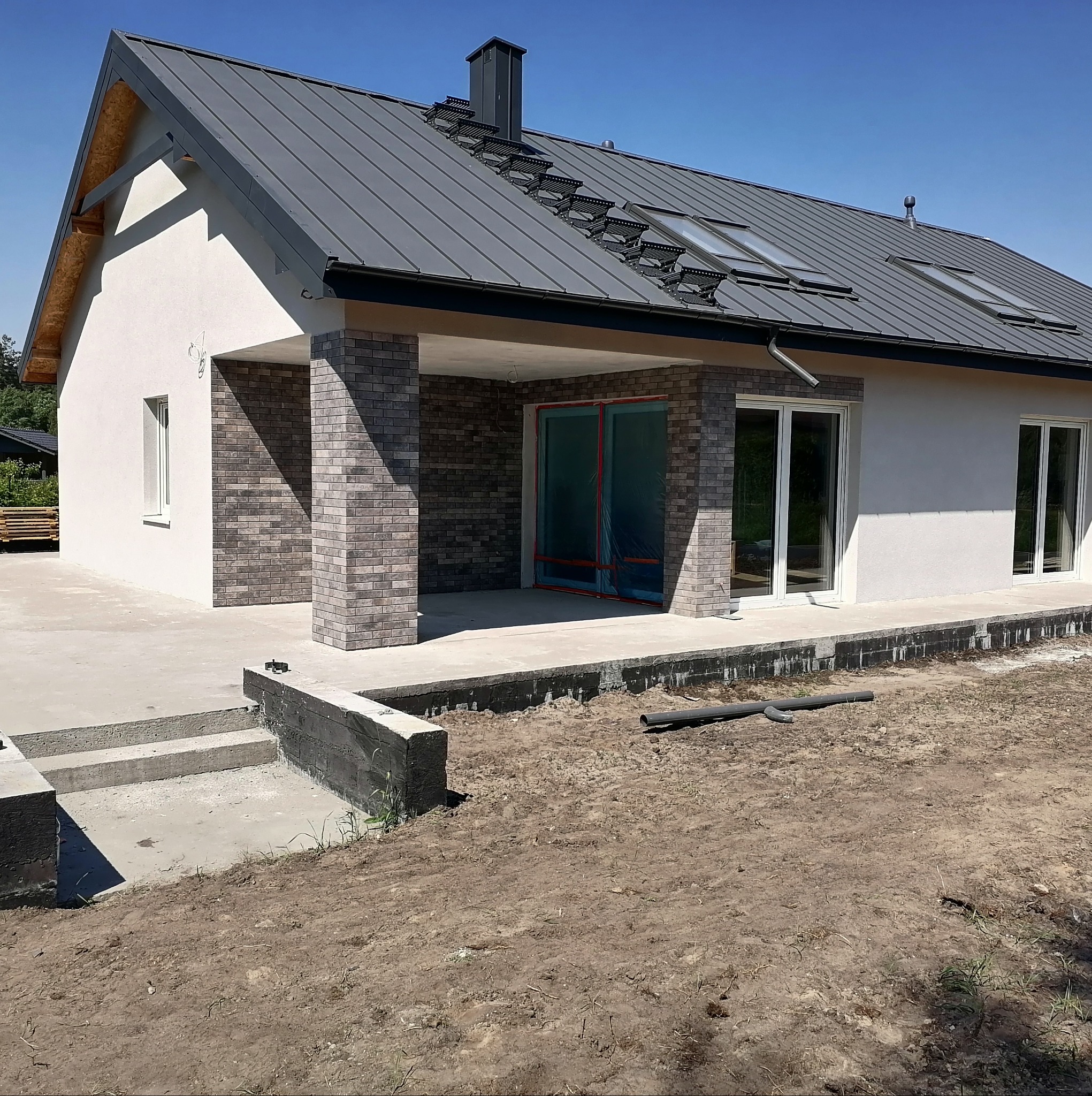 Nowoczesny dom jednorodzinny z szarą blachodachówką i elewacją w odcieniach bieli i szarości. Widoczne okna dachowe, ceglane filary i przygotowany teren wokół budynku.