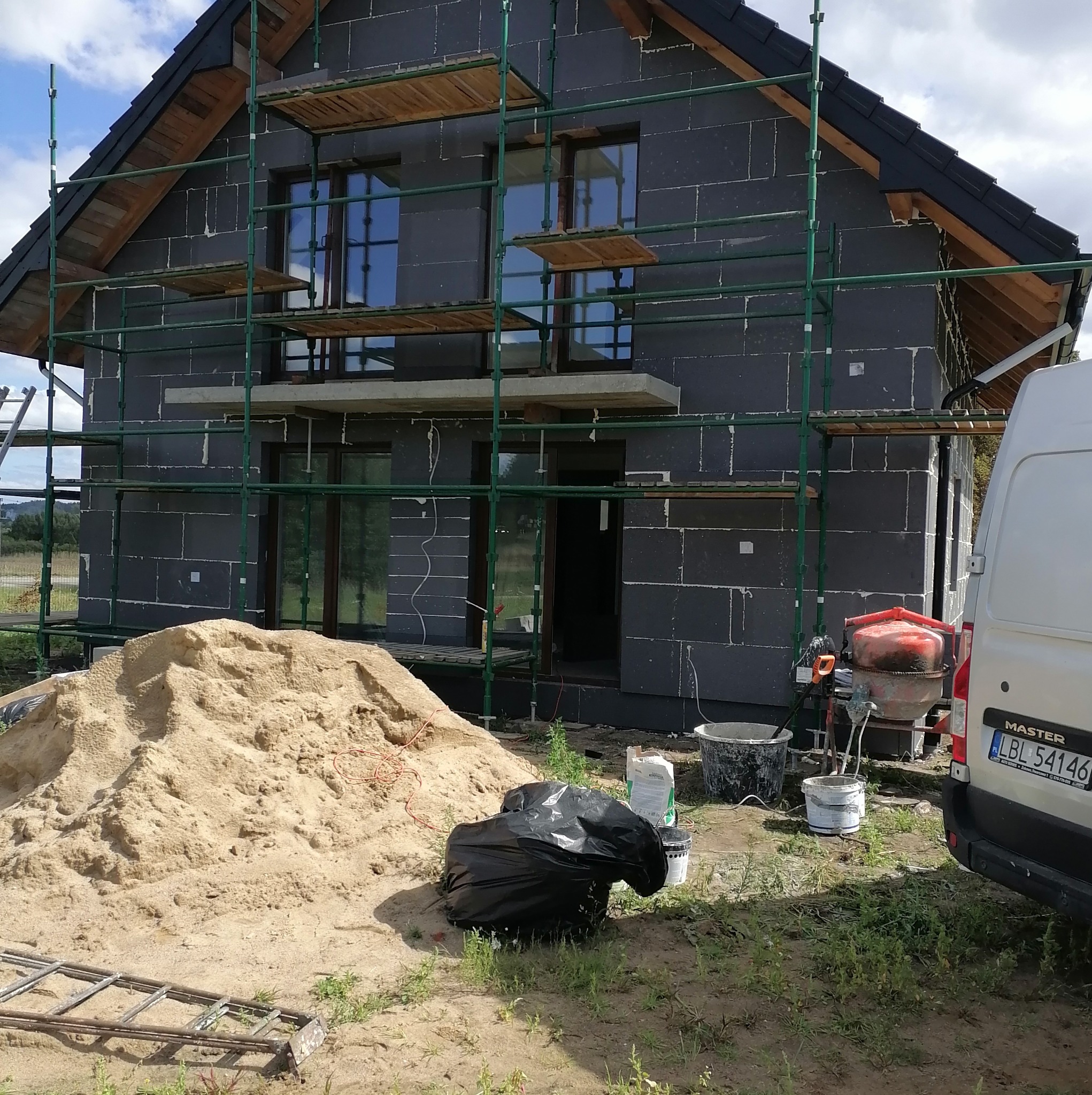 Dom w trakcie ocieplania, widoczne rusztowania, bloczki z szarego betonu komórkowego, piasek, betoniarka i samochód dostawczy. Prace elewacyjne w toku.