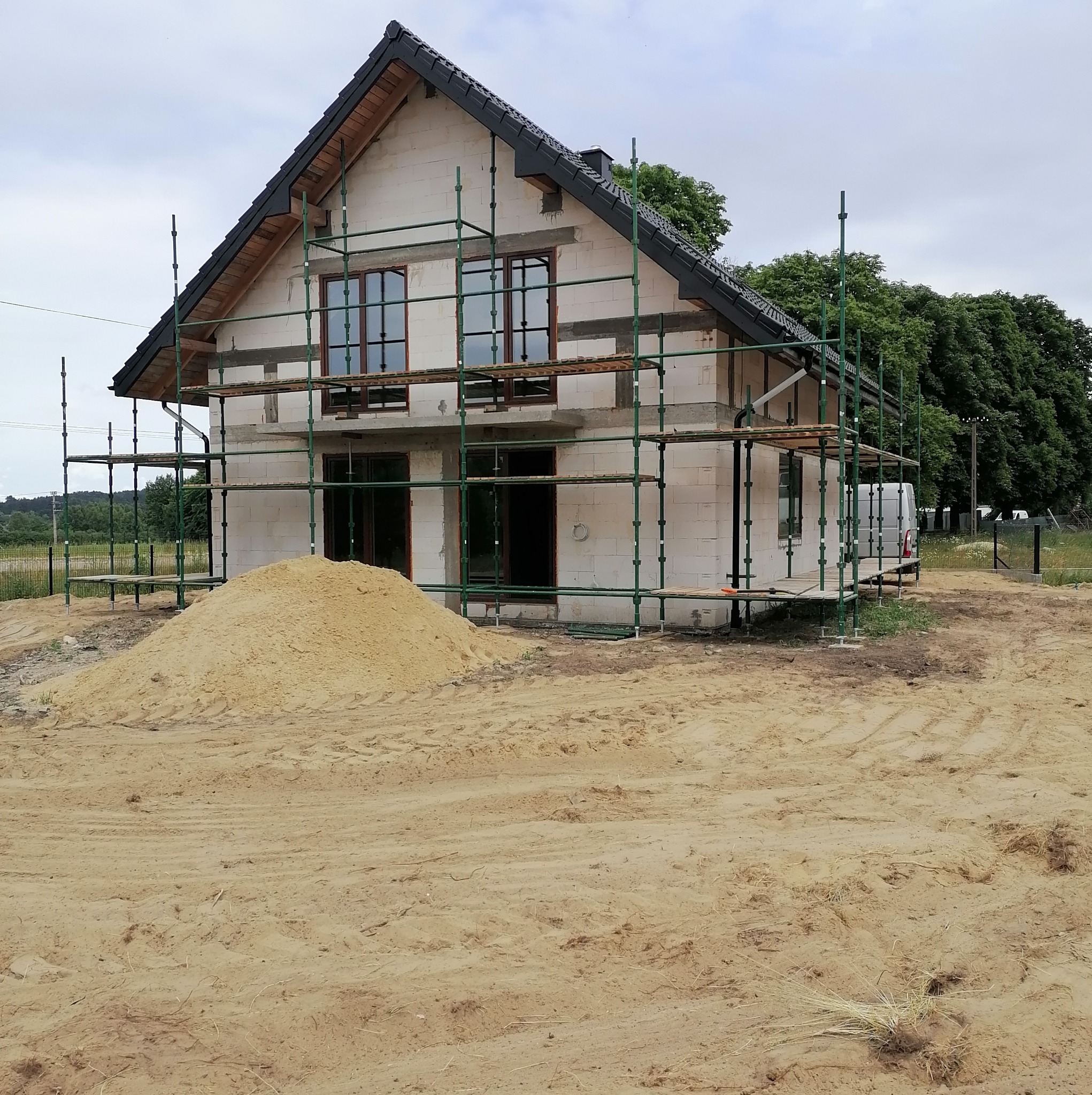 Niedokończony dom z białych bloczków, otoczony rusztowaniem i stertą piasku. Widoczne okna z brązowymi ramami i czarny dach. W tle zieleń drzew i częściowo widoczny biały samochód.