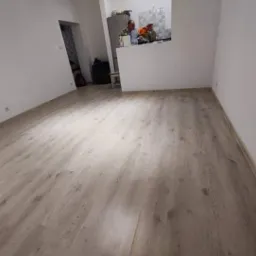 Jasnoszara podłoga z paneli laminowanych w nowym pomieszczeniu, widoczne niedoskonałości przy łączeniu z białą ścianą, w tle otwarta kuchnia z widoczną osobą.