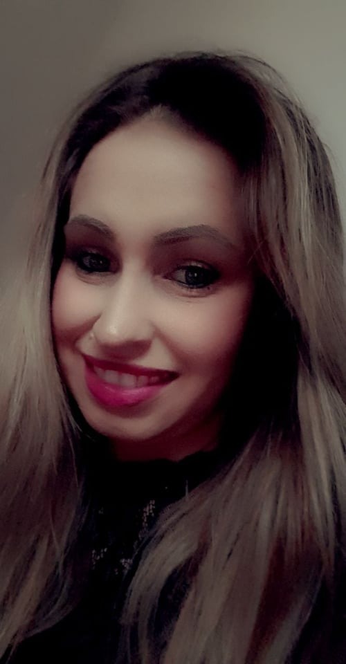 Portret uśmiechniętej kobiety z kolczykiem w nosie, pomalowanymi ustami w kolorze fuksji i blond włosami z ciemnymi odrostami.