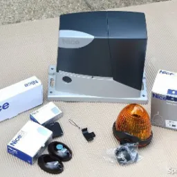 Zestaw do automatyki bramy przesuwnej marki Nice, zawierający napęd, lampę ostrzegawczą, piloty i akcesoria, rozłożony na beżowym tle.