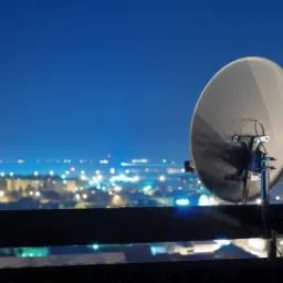 Antena satelitarna zamontowana na dachu budynku z widokiem na nocne miasto, rozświetlone licznymi światłami.