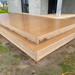 Nowoczesny taras z desek kompozytowych z dwoma stopniami, widoczny kabel elektryczny na ziemi, tło stanowi dom w trakcie budowy.