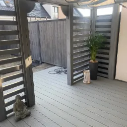 Szara pergola z ażurowymi ściankami i posadzką z desek w Ząbkowicach Śląskich, dekorowana figurką Buddy i doniczką z zieloną rośliną. W tle widoczne ogrodzenie.