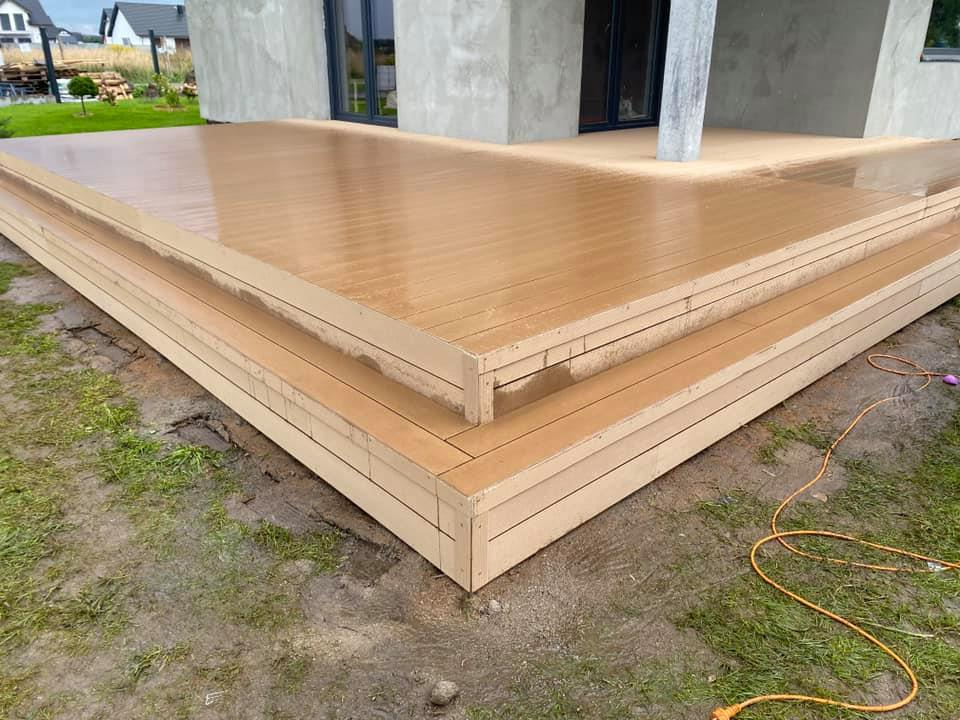 Nowoczesny taras z desek kompozytowych z dwoma stopniami, widoczny kabel elektryczny na ziemi, tło stanowi dom w trakcie budowy.