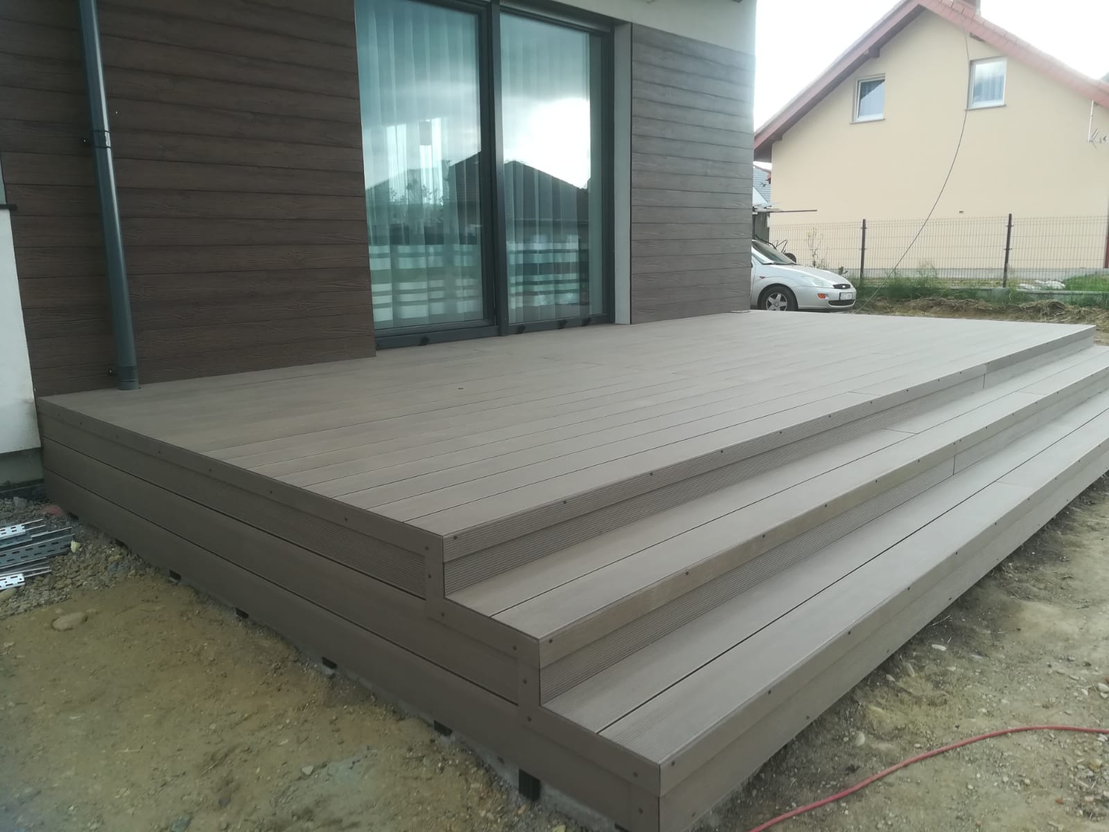 Nowoczesny taras z kompozytu w odcieniach szarości, z trzema stopniami, przylegający do domu z brązową elewacją i dużymi przesuwnymi oknami. W tle widoczny dom jednorodzinny i zaparkowany samochód.