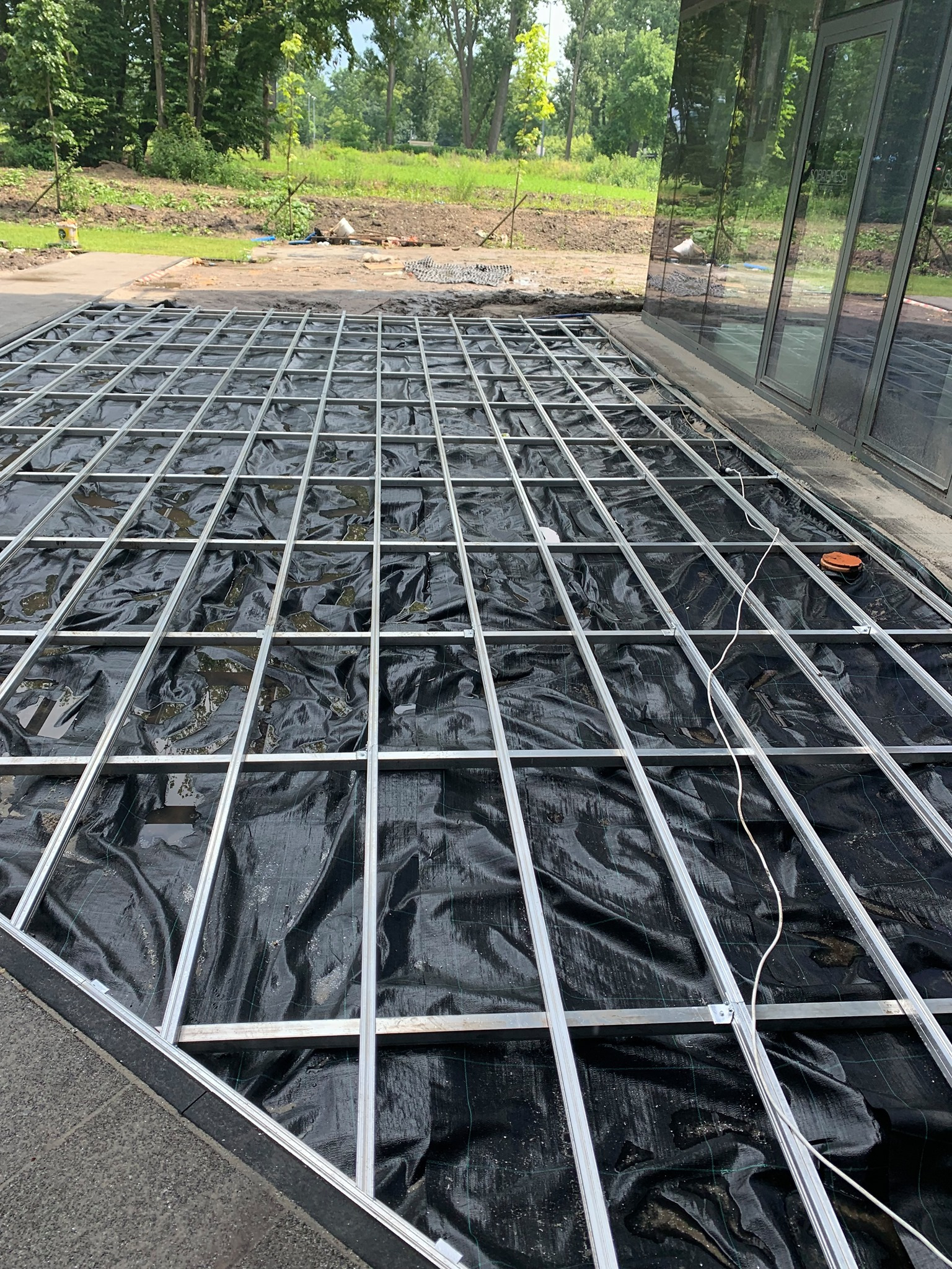 Metalowa konstrukcja nośna na czarnej geowłókninie, przygotowana pod montaż desek tarasowych, widoczna na tle zieleni i budynku z lustrzanymi szybami.