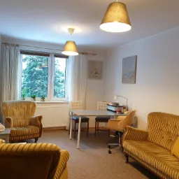 Jasne wnętrze gabinetu z fotelami w stylu retro, stołem i widokiem na zieleń za oknem.