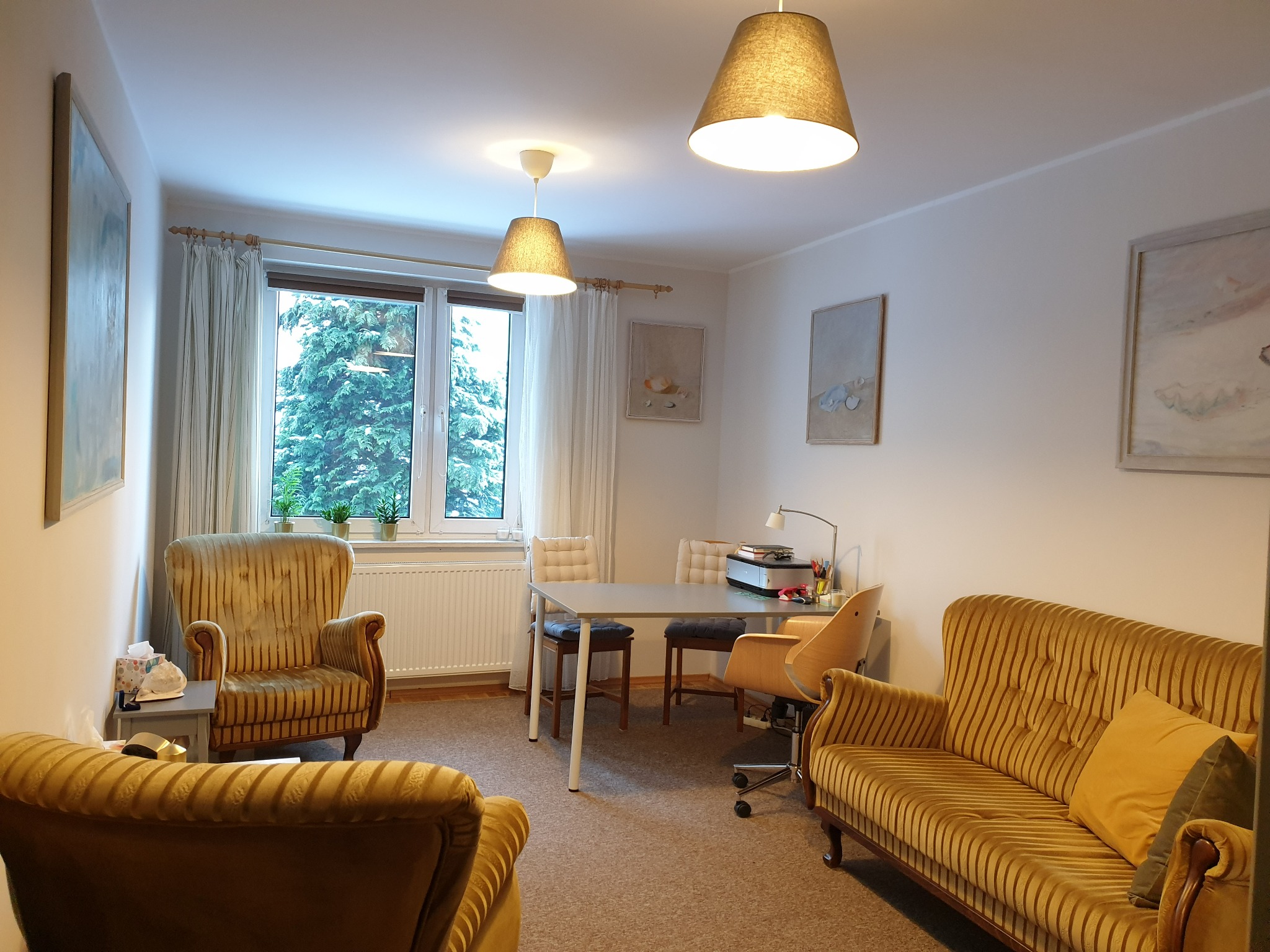 Jasne wnętrze gabinetu z fotelami w stylu retro, stołem i widokiem na zieleń za oknem.