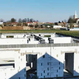 Stan surowy otwarty domu jednorodzinnego z białego bloczka, widok z góry na tle wiejskiej zabudowy i kościoła.