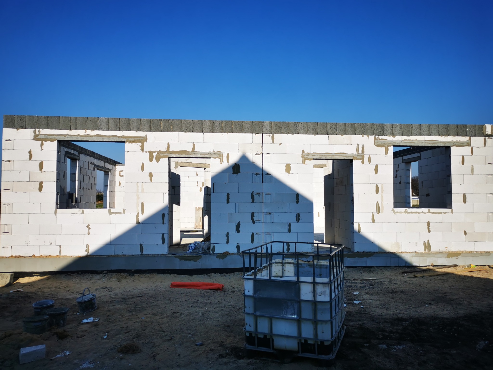 Wznoszone ściany domu z betonu komórkowego Ytong, widoczne otwory okienne i drzwiowe, górna warstwa z szarego betonu, w tle intensywnie błękitne niebo, na pierwszym planie paletopojemnik i wiaderka...