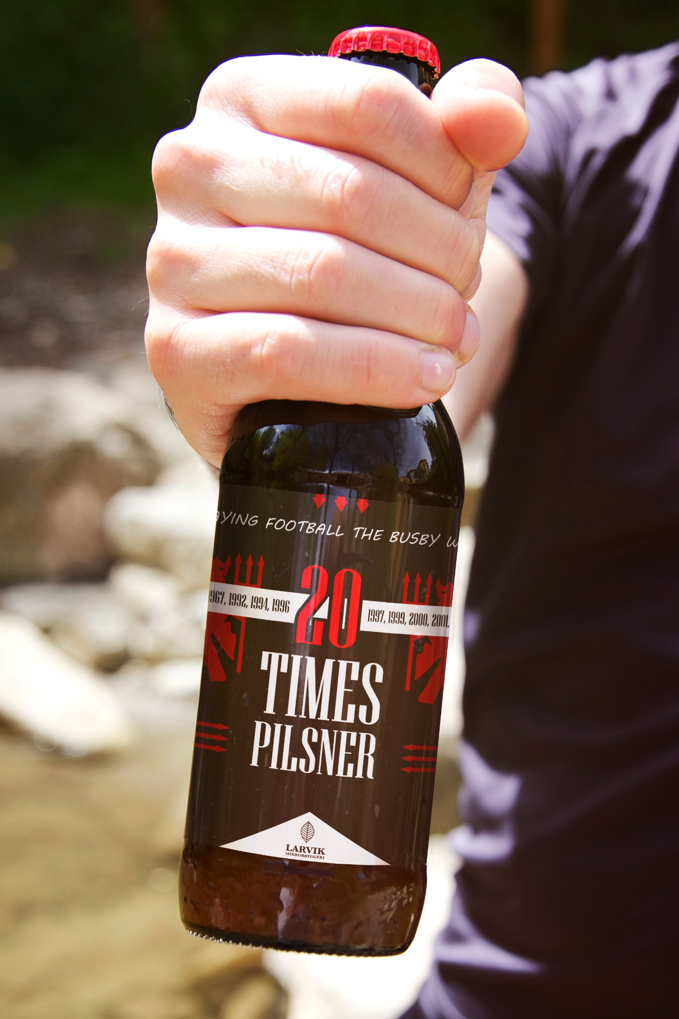 Męska dłoń trzyma butelkę ciemnego piwa rzemieślniczego z etykietą '20 Times Pilsner', na tle rozmytego krajobrazu z elementami roślinności i kamieni.