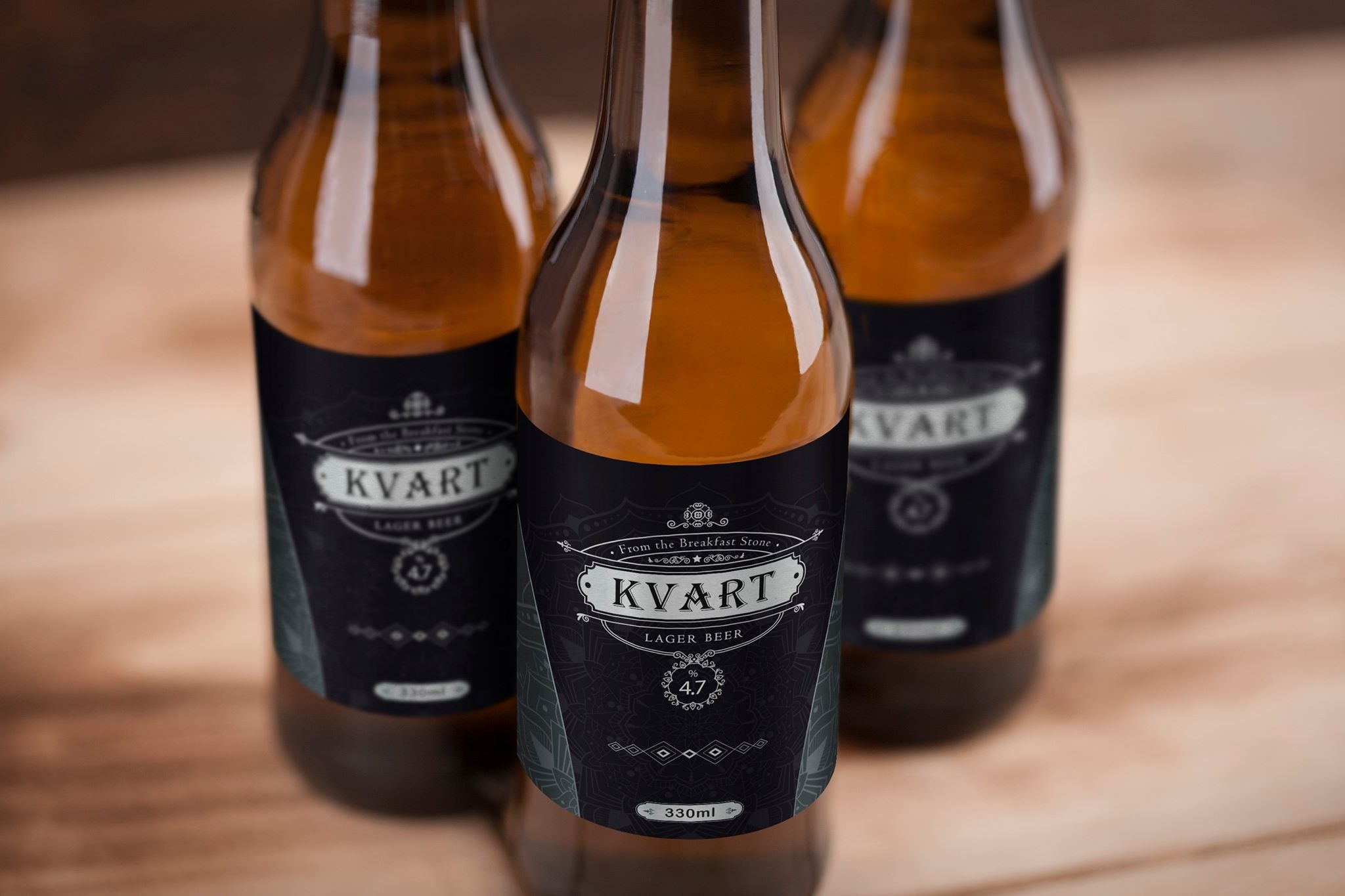 Trzy butelki bursztynowego lagera Kvart z etykietą w stylu vintage na drewnianym stole, prezentujące projekt graficzny identyfikacji wizualnej.