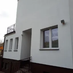 Odnowiona elewacja budynku z białym tynkiem strukturalnym, brązowym cokołem i metalowymi balustradami, widoczna lampa elewacyjna i okna z odbiciem nieba.