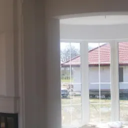 Wnętrze domu w trakcie malowania, widoczne białe ściany, okno z widokiem na dom z czerwoną dachówką i fragment kominka.