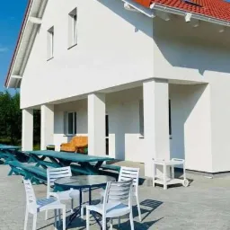 Biały, dwukondygnacyjny dom z czerwonym dachem, widok z zewnątrz, z tarasem i meblami ogrodowymi.