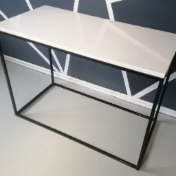 Minimalistyczny stolik konsola z białym blatem i czarną, stalową ramą, wykonany w Strzyżowicach, na tle ściany z geometrycznym wzorem.