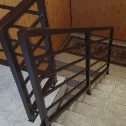 Metalowa balustrada schodowa w kolorze ciemnego brązu, z prostymi, geometrycznymi elementami, zamocowana do jasnych, kafelkowych schodów wewnątrz budynku, widok z góry.