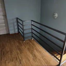 Metalowa balustrada schodowa z poziomych prętów, zamontowana w nowoczesnym wnętrzu z drewnopodobną podłogą i szarą ścianą. Widoczny fragment schodów z drewnianymi stopniami.
