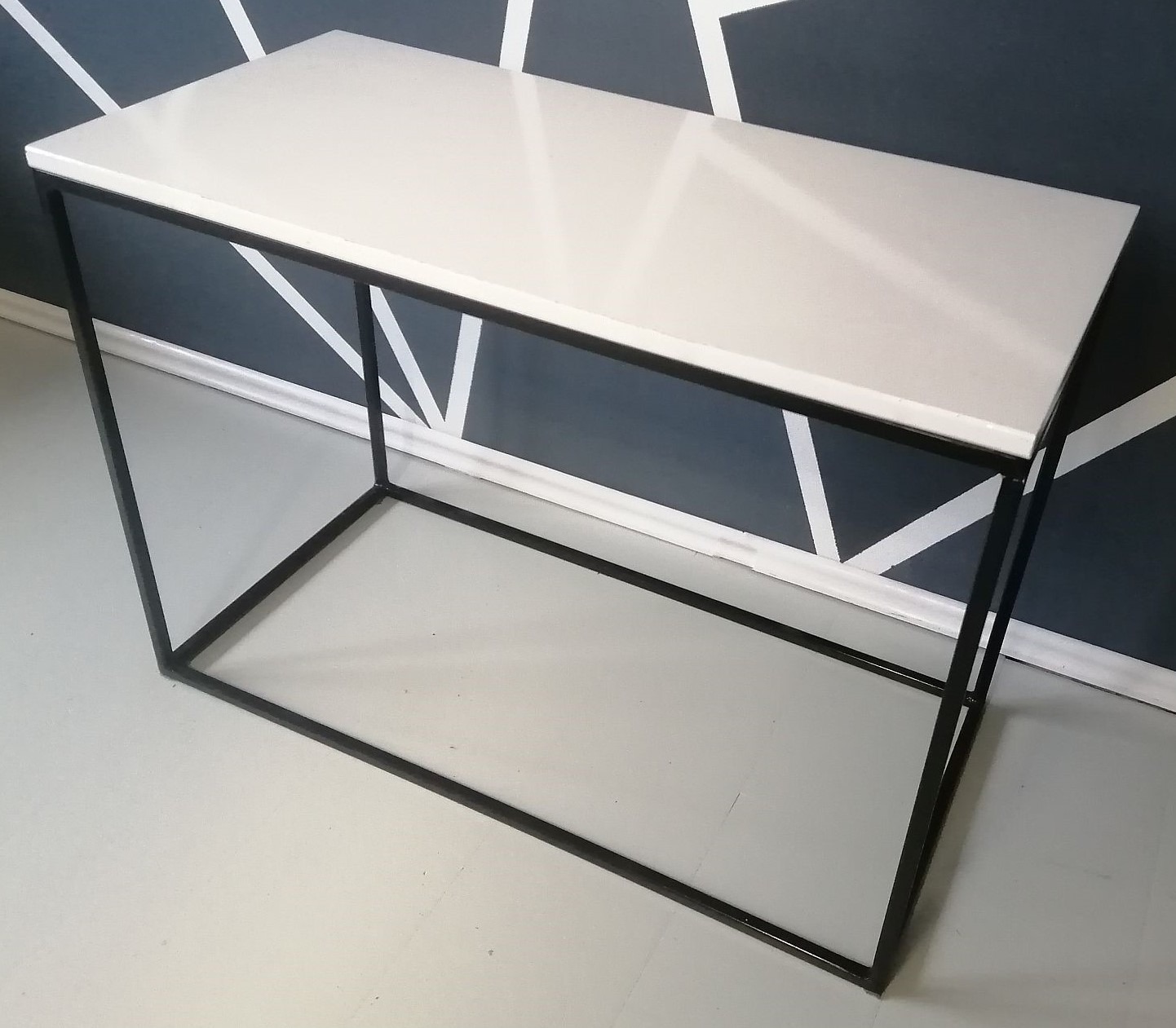 Minimalistyczny stolik konsola z białym blatem i czarną, stalową ramą, wykonany w Strzyżowicach, na tle ściany z geometrycznym wzorem.