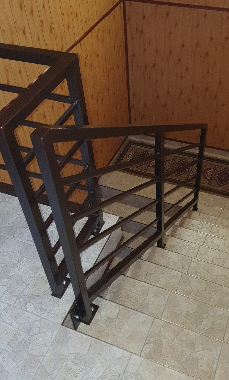 Metalowa balustrada schodowa w kolorze ciemnego brązu, z prostymi, geometrycznymi elementami, zamocowana do jasnych, kafelkowych schodów wewnątrz budynku, widok z góry.