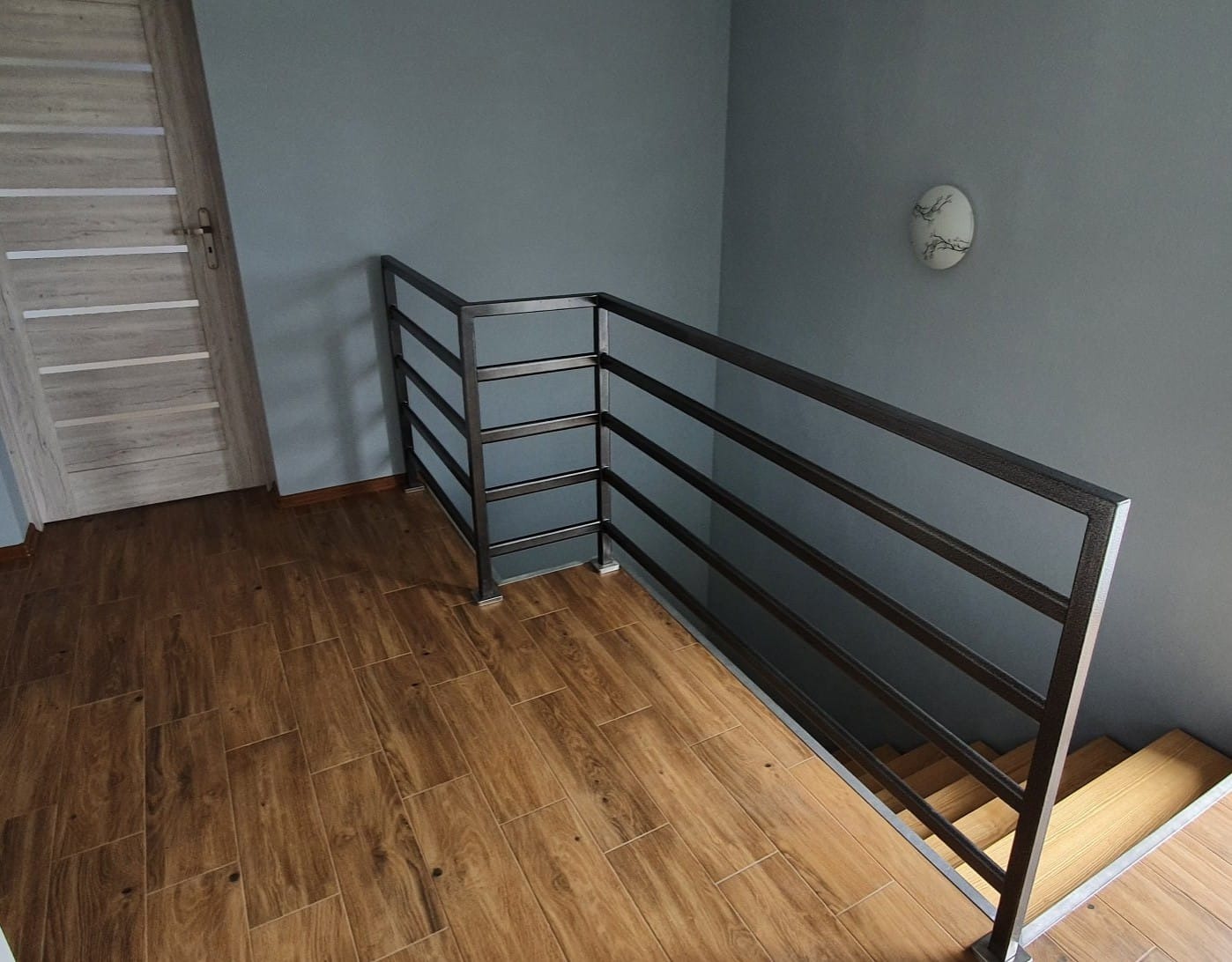Metalowa balustrada schodowa z poziomych prętów, zamontowana w nowoczesnym wnętrzu z drewnopodobną podłogą i szarą ścianą. Widoczny fragment schodów z drewnianymi stopniami.