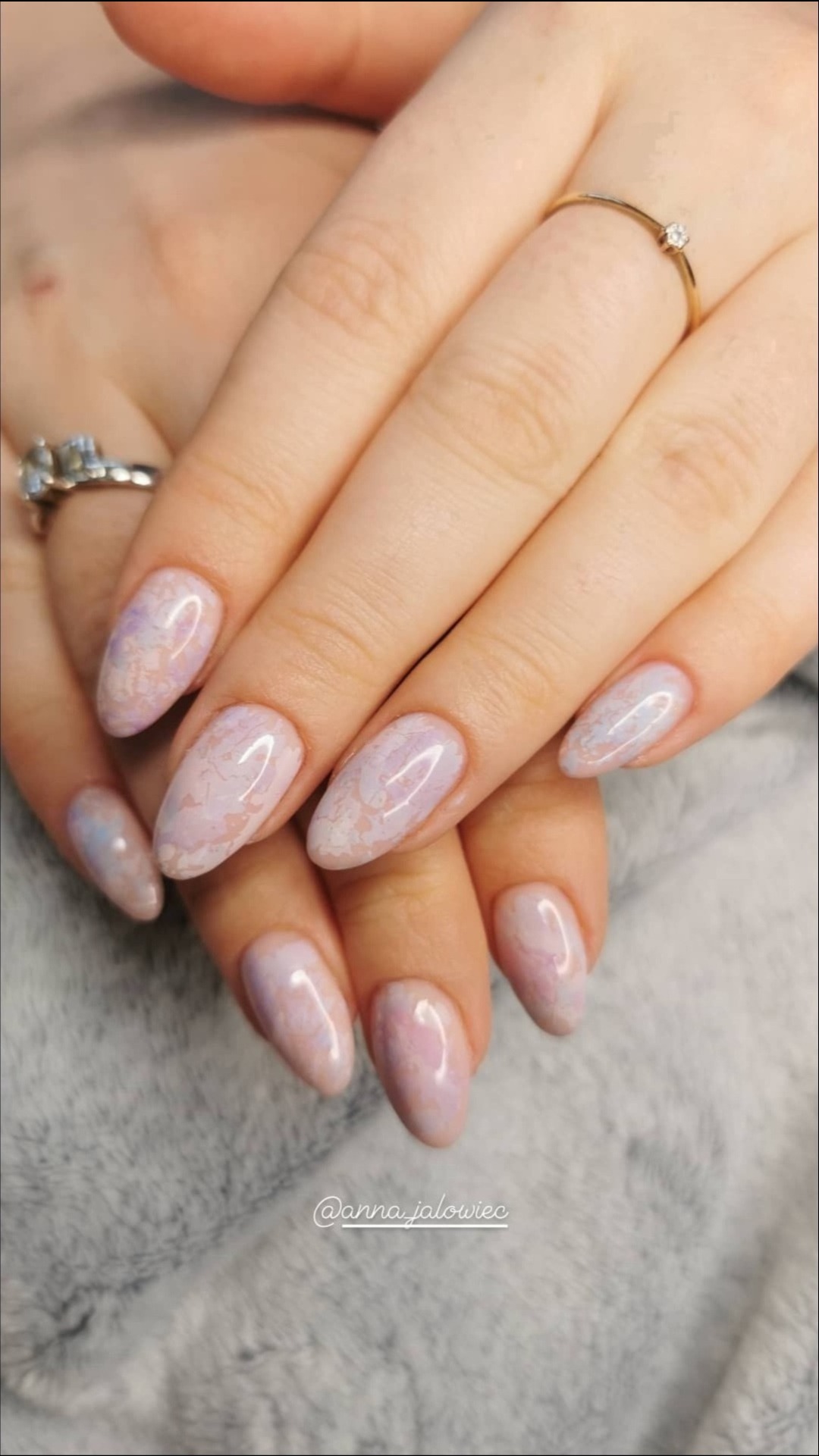 Dłonie kobiety z migdałowymi paznokciami w delikatnym marmurkowym wzorze w odcieniach bieli i fioletu, ozdobione złotym pierścionkiem z małym kamieniem i srebrnym pierścionkiem z większym kamieniem...