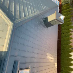 Nowy, szary dach panelowy z oknami dachowymi i kominkiem wentylacyjnym, widok z góry, w tle tuje i materiały budowlane.