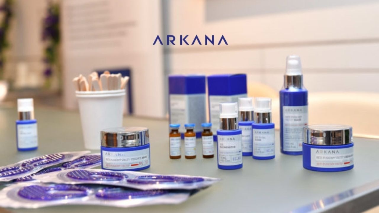 Zestaw kosmetyków i dermokosmetyków marki ARKANA: kremy, serum, maski w niebiesko-srebrnych opakowaniach, widoczne logo firmy, w tle kubek z drewnianymi szpatułkami.