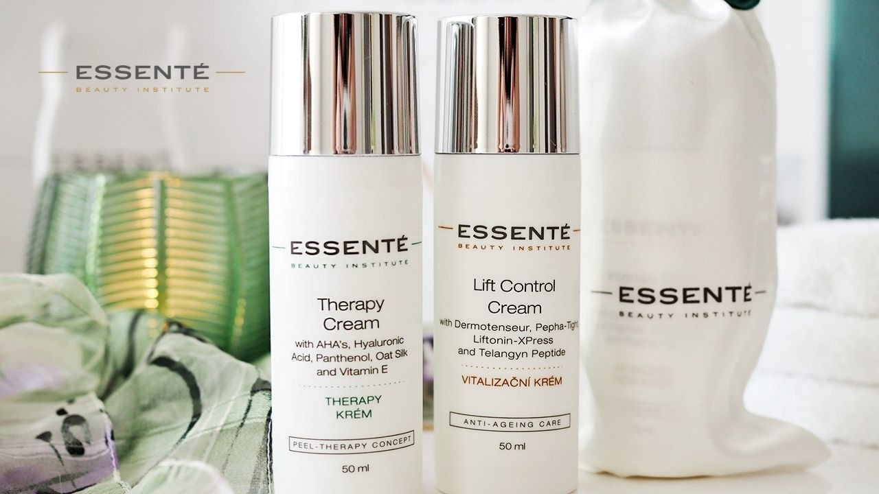 Dwa białe flakony kremów ESSENTÉ z srebrnymi nakrętkami: Therapy Cream z kwasami AHA i Lift Control Cream z Dermotenseur, na jasnym tle z zielonym akcentem dekoracyjnym i białymi ręcznikami.