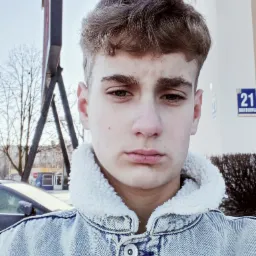 Młody mężczyzna w dżinsowej kurtce z futrzanym kołnierzem, na tle budynku z numerem 21 i drzew bez liści.